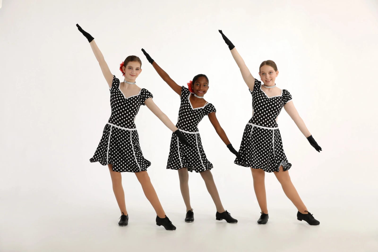 Three tap dancers in black and white polka dot dresses on the white studio backdrop.