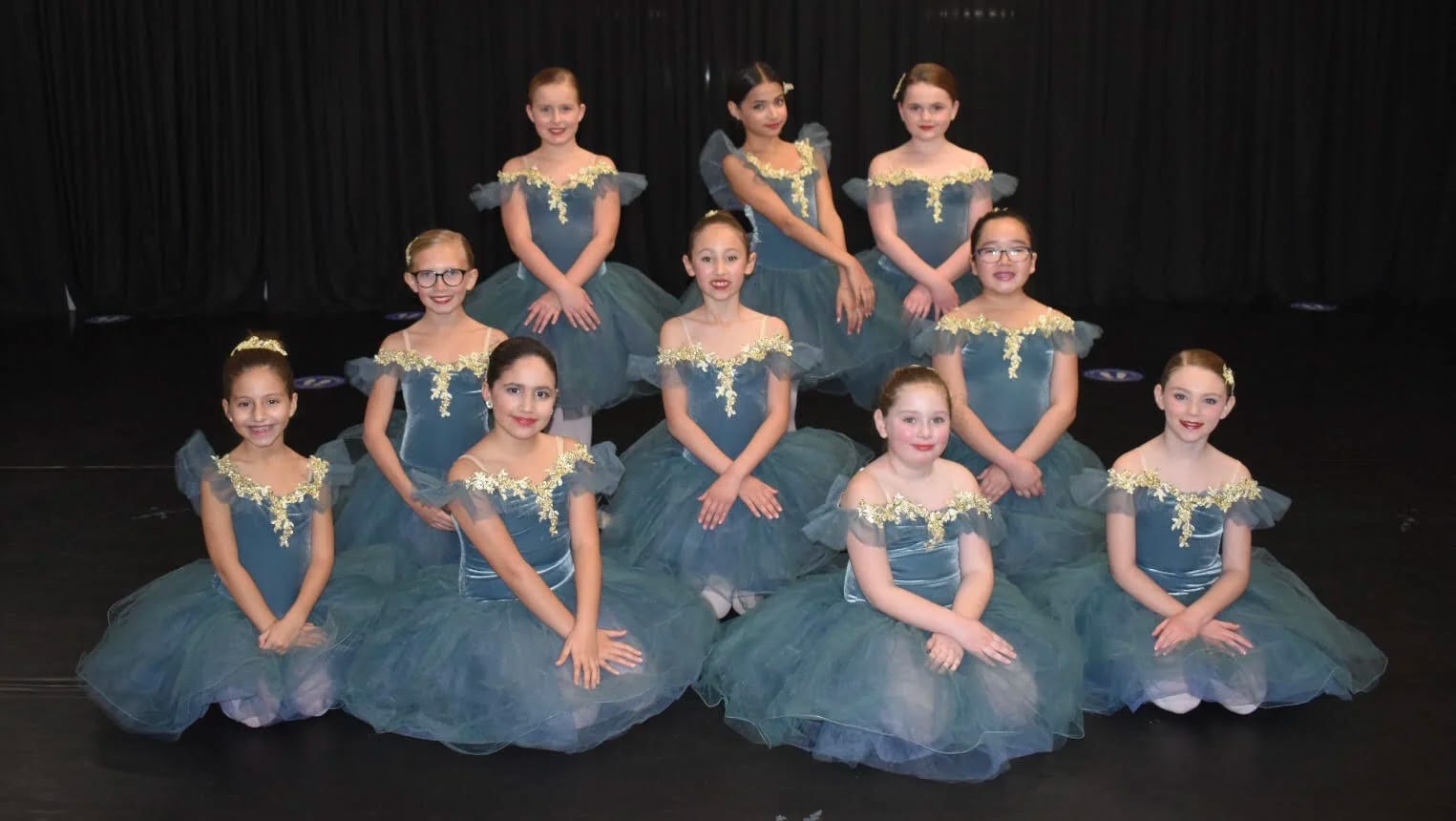 A ballet group in long teal tutus seated and standing on a dark stage.