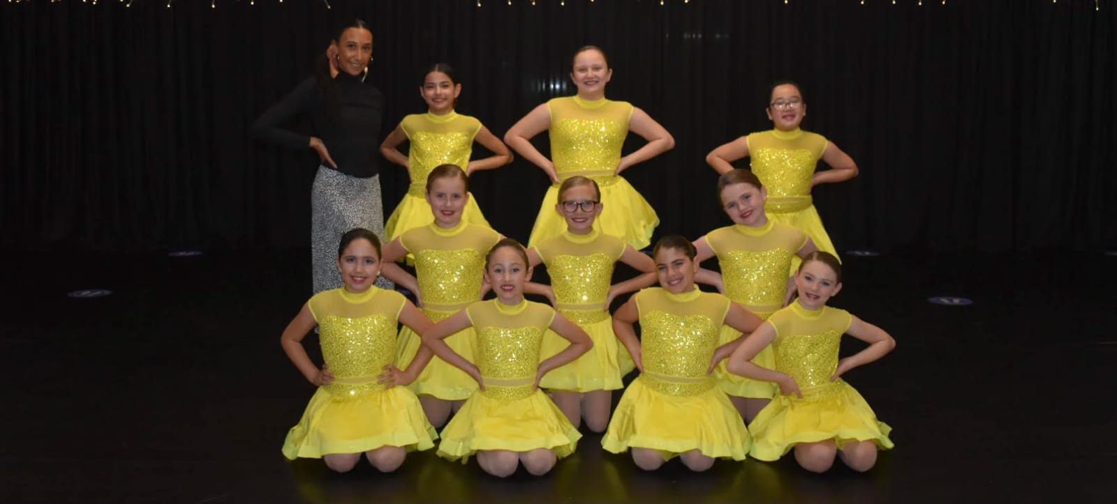 A jazz group in gold sequin dresses kneeling on stage with one of our teachers.